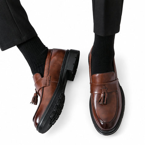 Sterling brown leather tassel loafers with a polished finish, moc toe detailing and sturdy black soles shown on a model for a discreet height boost