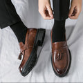 Close-up of men's brown leather tassel loafers named Sterling with black soles, worn with black socks and trousers, designed for discreet height increase and formal style.