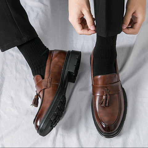 Close-up of men's brown leather tassel loafers named Sterling with black soles, worn with black socks and trousers, designed for discreet height increase and formal style.