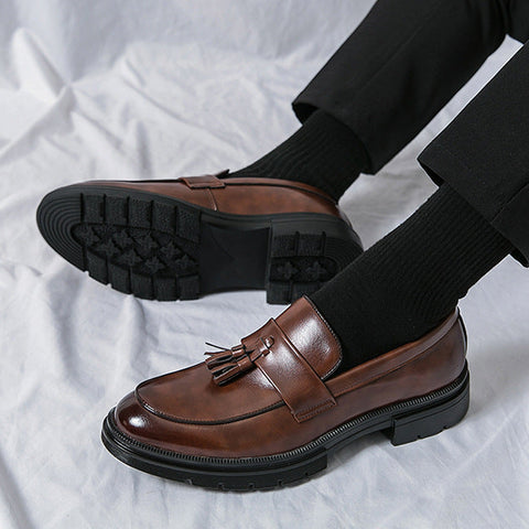 Pair of polished brown Sterling loafers with tassel detail and black rubber soles, worn with black socks and formal black trousers, designed for discreet height boost and comfort.