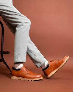 Man seated on a stool in cream trousers, one leg extended, wearing tan leather casual shoes with cream soles