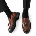 Sterling brown leather tassel loafers with a polished finish, moc toe detailing and sturdy black soles shown on a model for a discreet height boost
