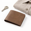 The Modern Wallet in brown leather with a bifold design displayed beside a metal key and beige buttoned shirt on a white background